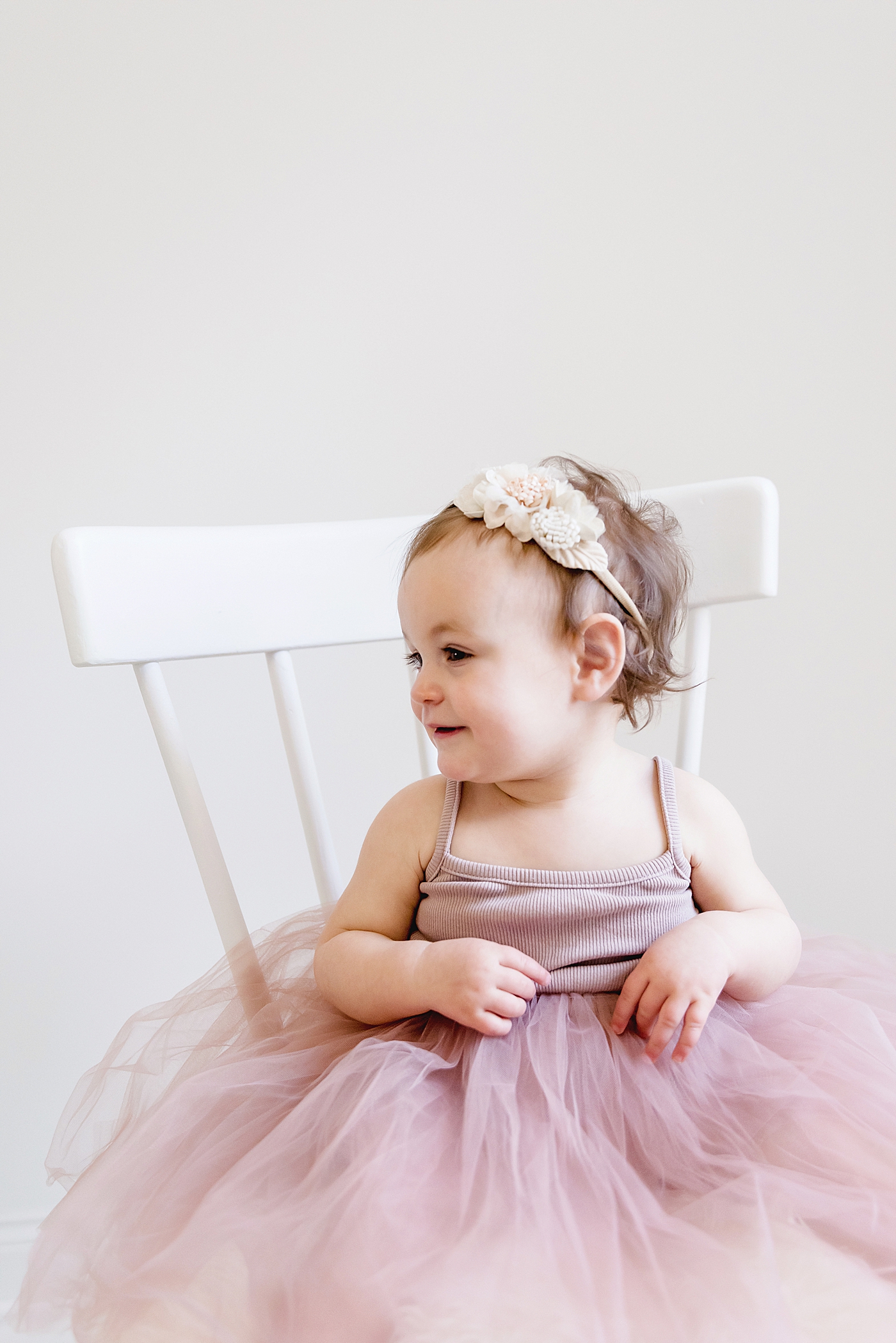 Baby girl in pink with floral bow sitting on white chair | Photo by Huntersville Baby Photographer Anna Wisjo 