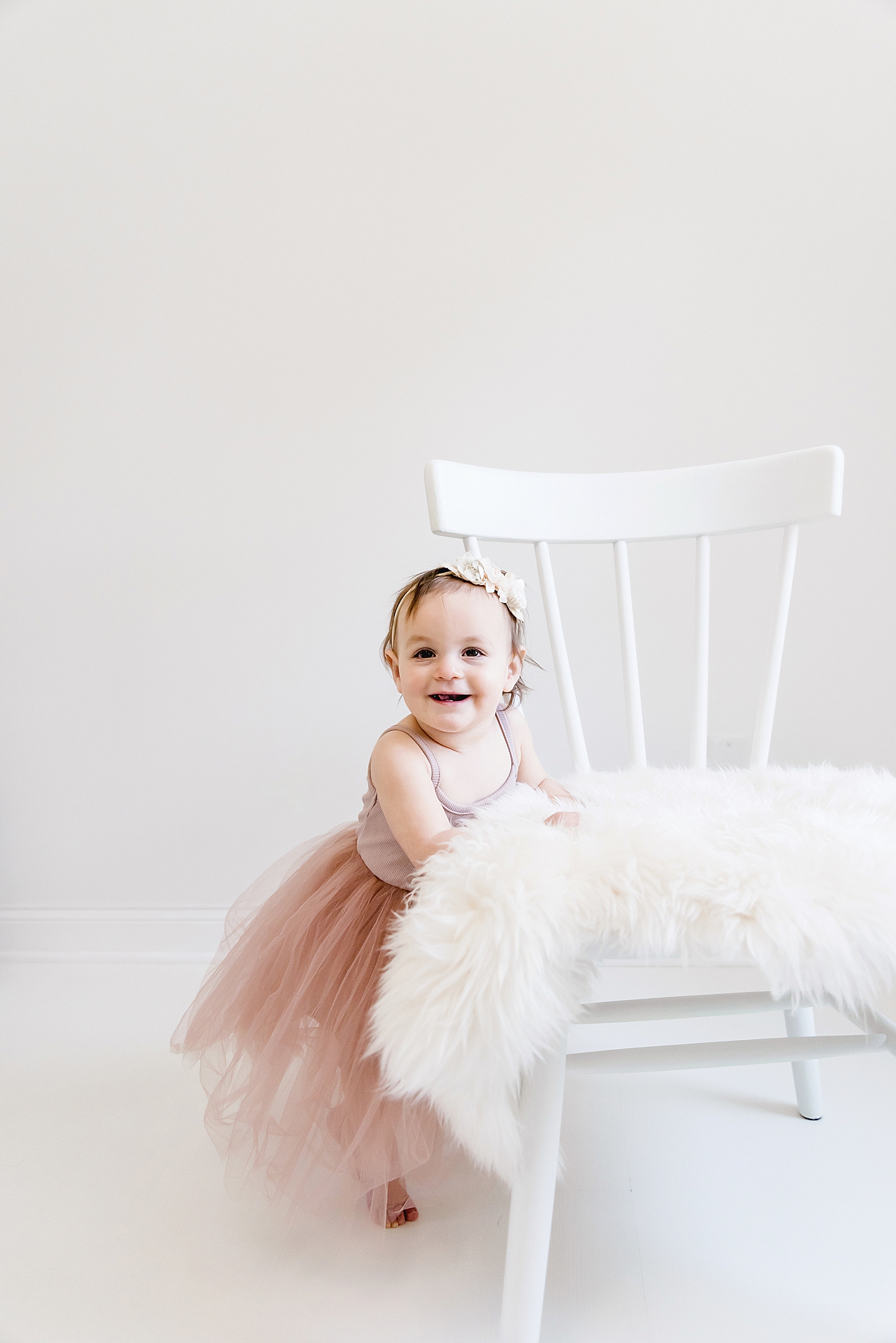 Baby girl in pink smiling at the camera standing near white chair | Photo by Anna Wisjo Photography
