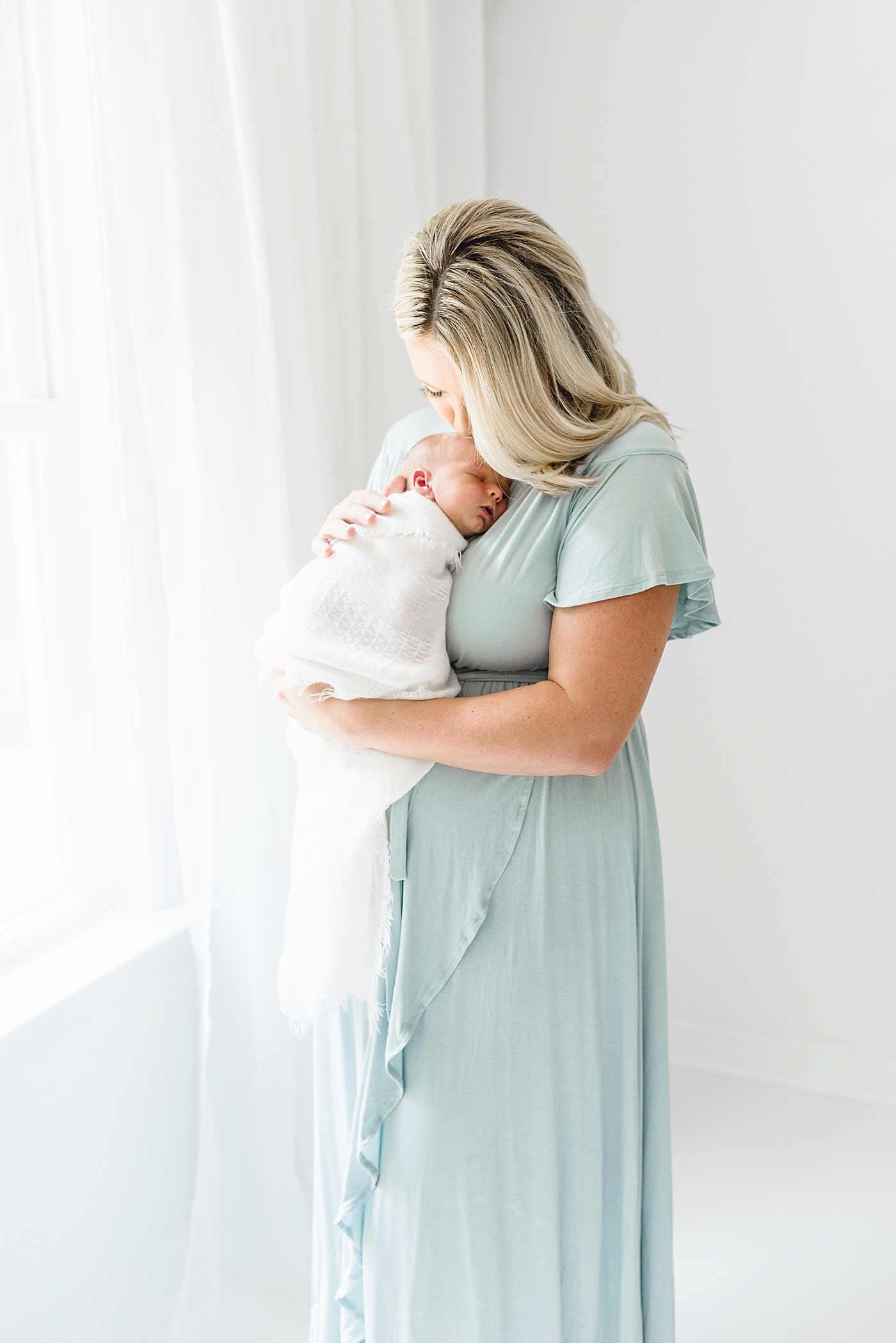 Mom in blue dress kissing newborn wrapped in white swaddle | Photo by Anna Wisjo Photography