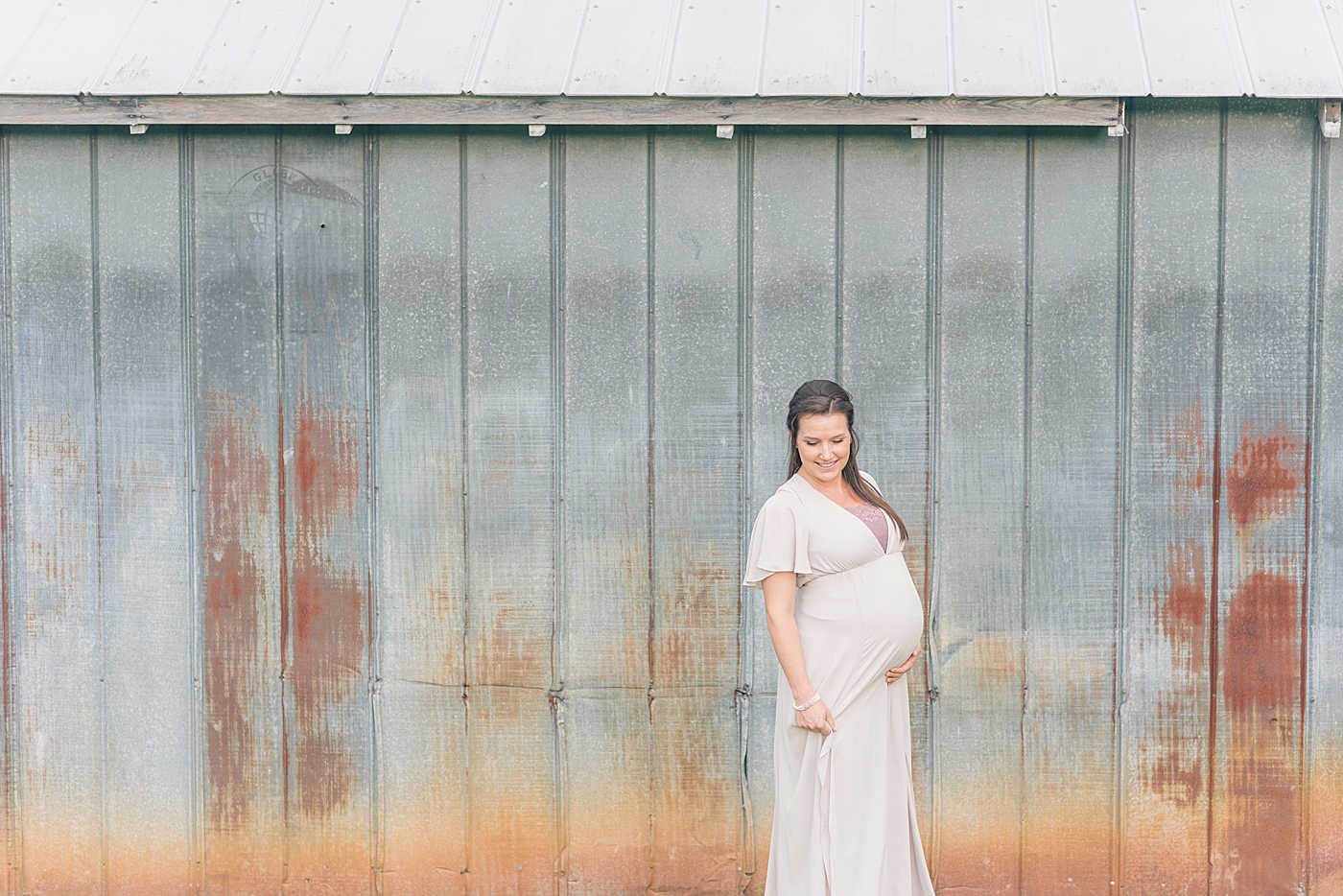 Mother to be in long white dress standing in front of a barn | Photo by Anna Wisjo Photography