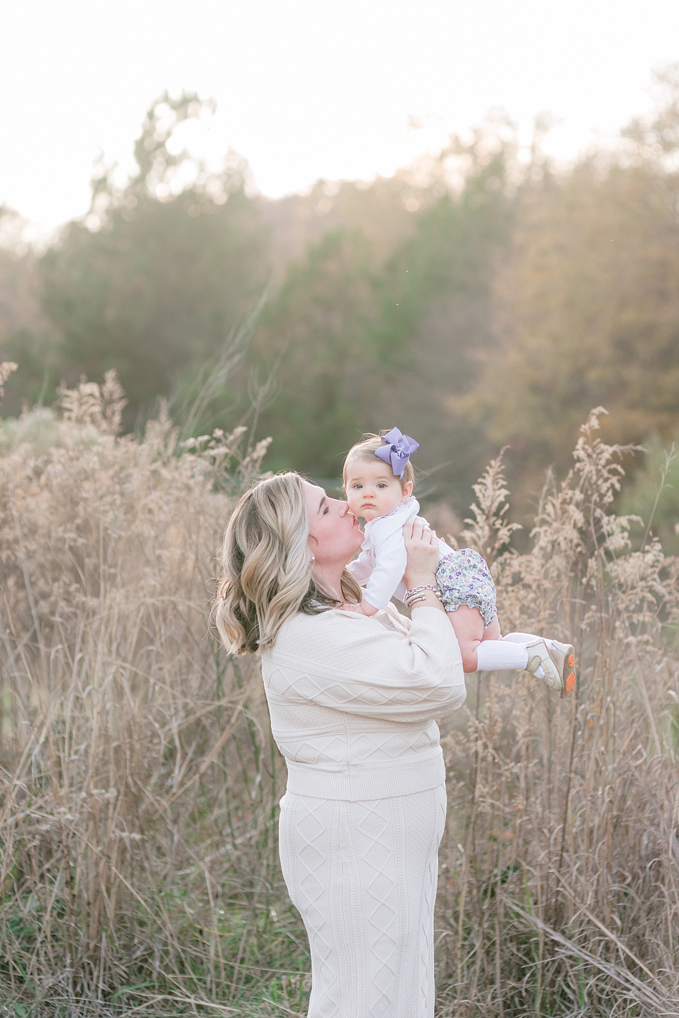 Mom kissing baby girl with purple bow during her Milestone Session at Marvin Efird Park | Photo by Anna Wisjo Photography 