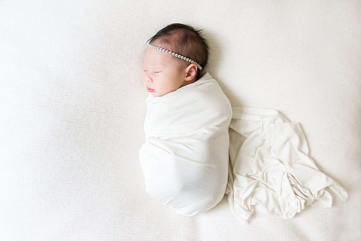 Newborn baby girl wrapped in a white swaddle with headband | Photo by Anna Wisjo Photography